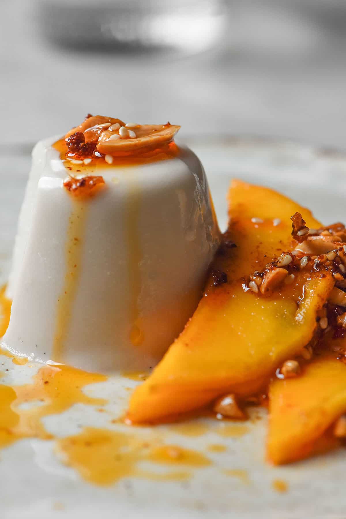 A brightly lit side on view of a coconut panna cotta topped with fresh mango and chilli crisp. The panna cotta sits atop a white ceramic plate atop a white marble table and a glass of water sits in the background