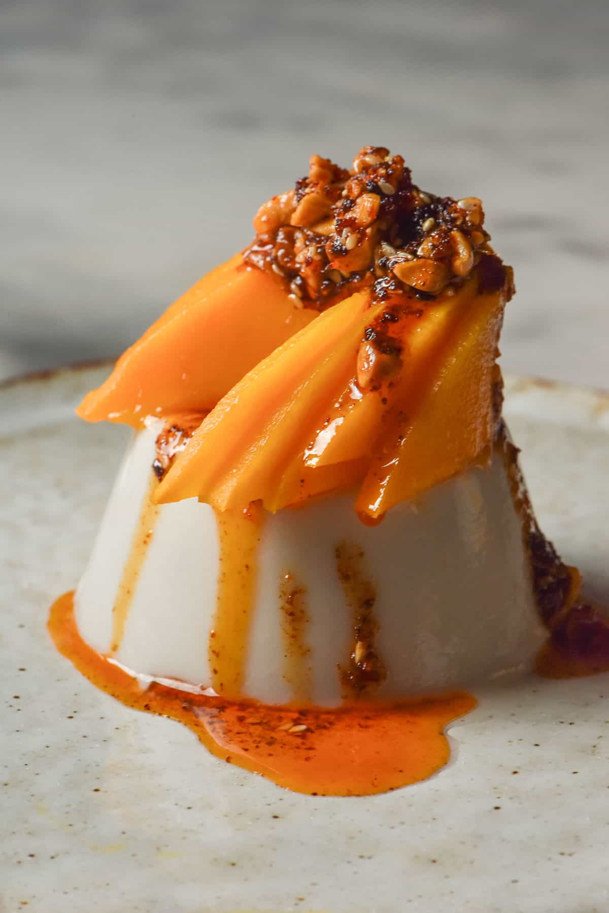 A side on image of a coconut panna cotta topped with mango and chilli crisp. The panna cotta sits on a white ceramic plate atop a white marble table 