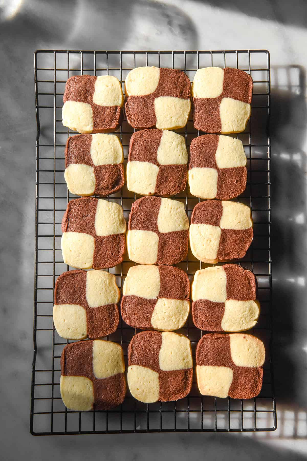 An aerial sunlit image of gluten free chocolate and vanilla checkerboard cookies on a cooling rack atop a white marble table