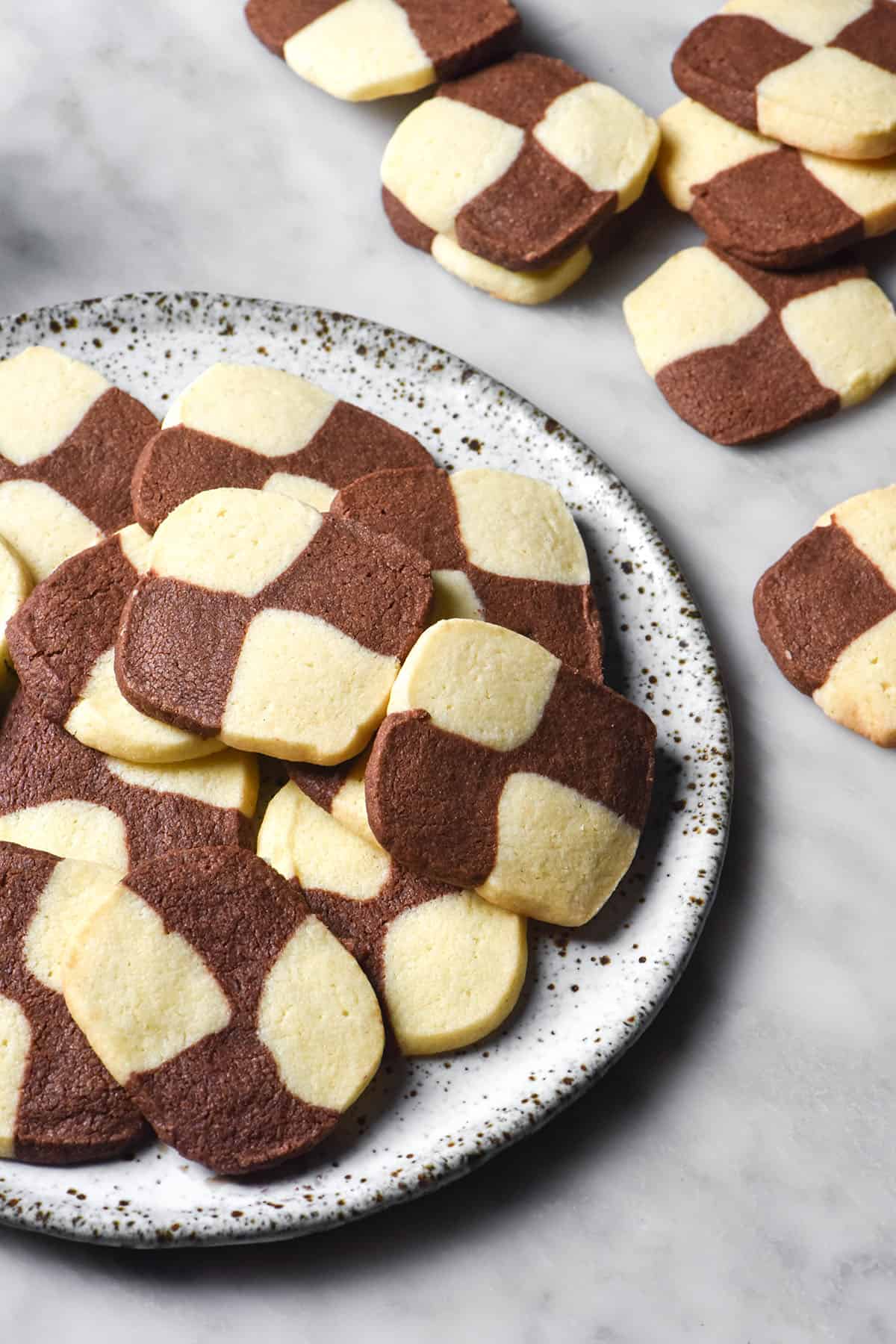 A side on image of a white speckled ceramic plate topped with gluten free checkerboard cookies on a white marble table. Extra checkerboard cookies sit on the table to the right of the plate