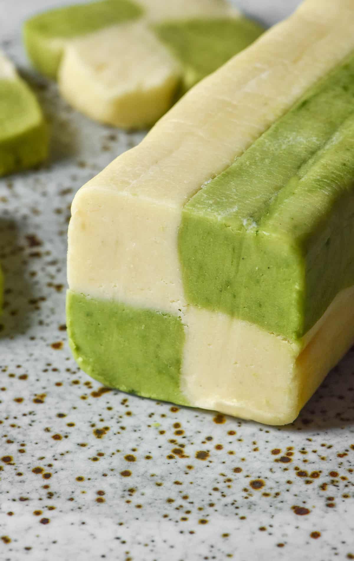 A close up side on image of a log of gluten free checkerboard cookie dough atop a white speckled ceramic plate. Some cookies have been sliced off the log and sit behind it on the plate