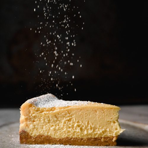 A moody side on image of a slice of gluten free cheesecake on a white plate and marble table against a dark backdrop. A sprinkle of icing sugar falls from the top of the image