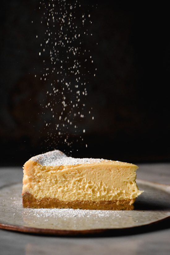 A moody side on image of a slice of gluten free cheesecake on a white plate and marble table against a dark backdrop. A sprinkle of icing sugar falls from the top of the image