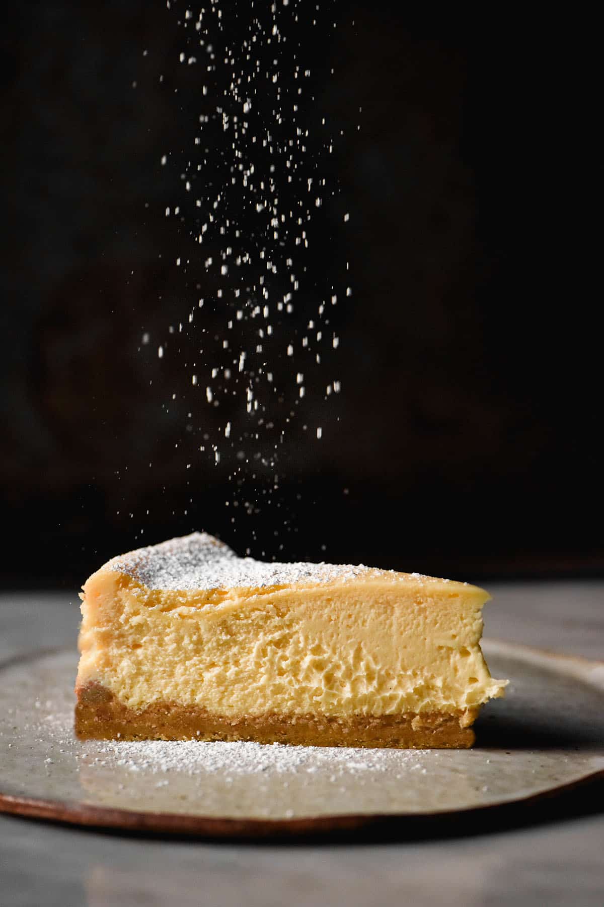 A moody side on image of a slice of gluten free cheesecake against a dark backdrop being sprinkled with icing sugar