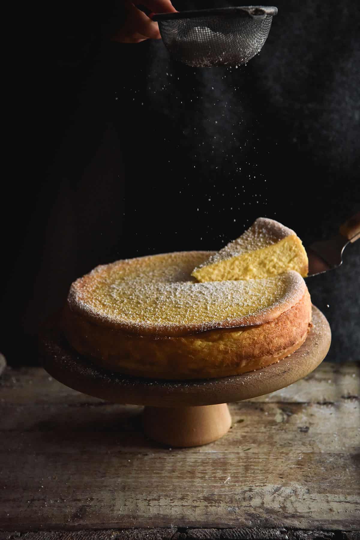 A moody image of a gluten free cheesecake on a cake stand being sprinkled with icing sugar