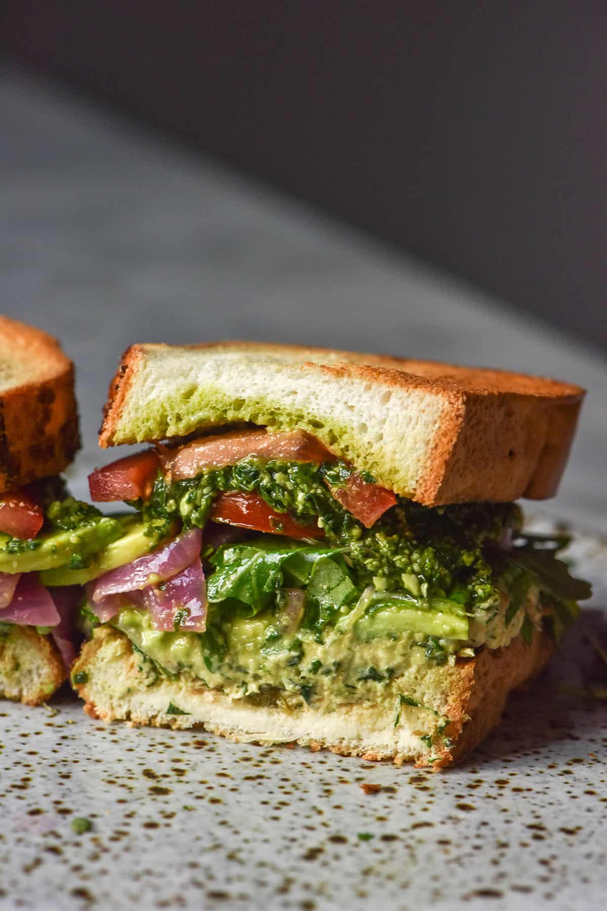 A side on close up image of half of a gluten free low FODMAP tunacado on a white marble table against a dark backdrop. The sandwich is filled with tuna mousse, rocket, pickled red onion, slices of tomato, avocado and low FODMAP pesto.