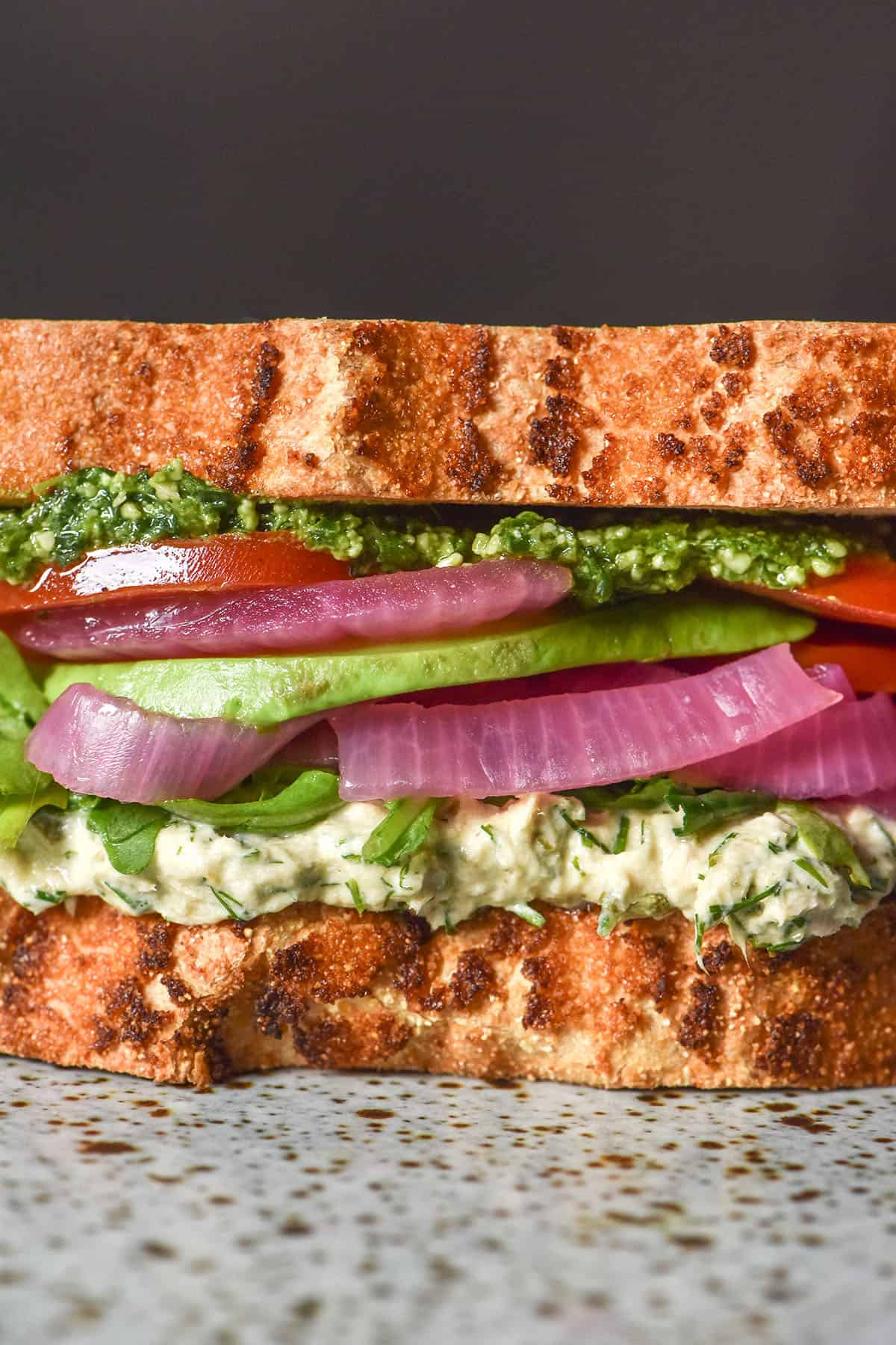 A side on close up image of a gluten free low FODMAP tunacado on a white speckled ceramic plate against a dark backdrop. The sandwich is filled with tuna mousse, rocket, pickled red onion, slices of tomato, avocado and low FODMAP pesto.