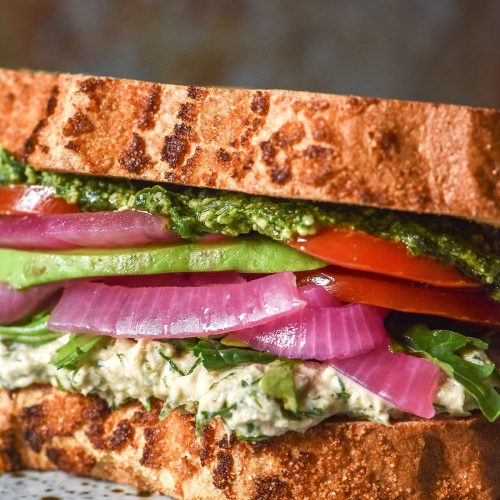 A side on close up image of a gluten free low FODMAP tunacado on a white speckled ceramic plate against a dark backdrop. The sandwich is filled with tuna mousse, rocket, pickled red onion, slices of tomato, avocado and low FODMAP pesto.