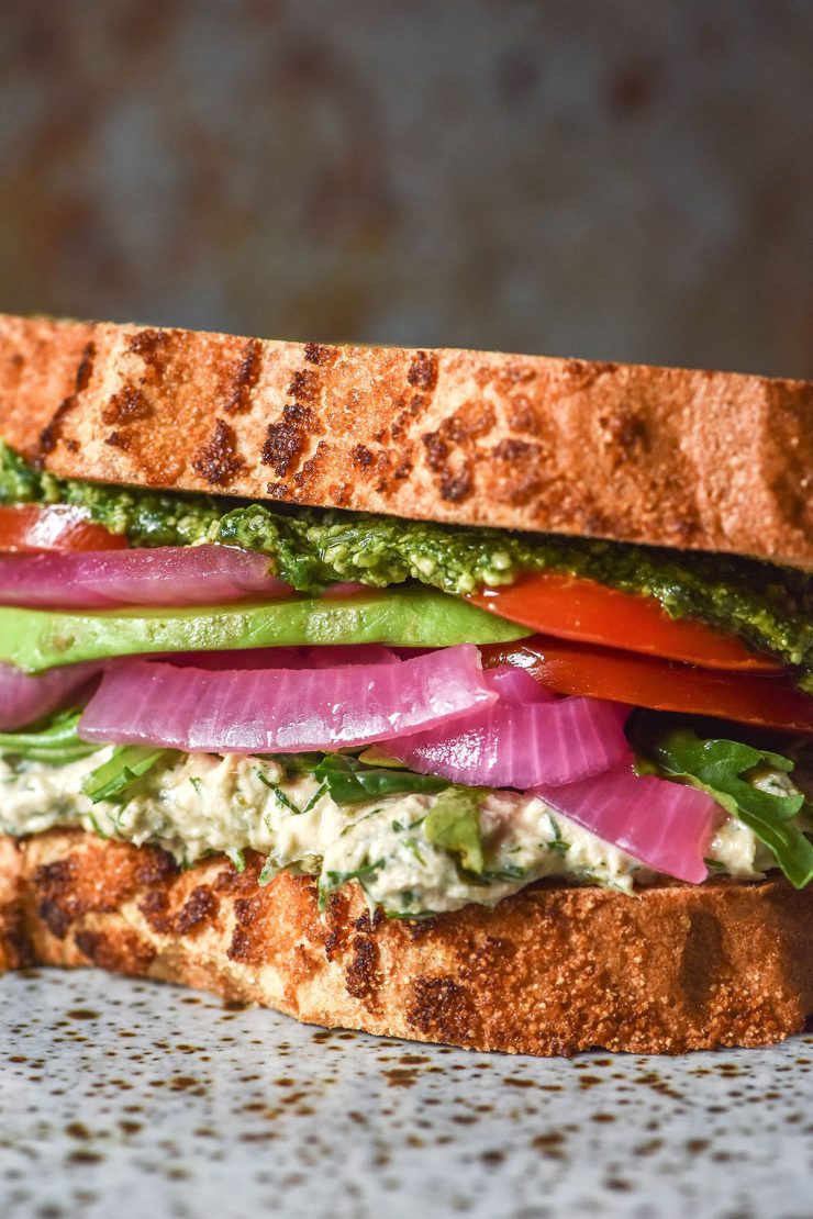 A side on close up image of a gluten free low FODMAP tunacado on a white speckled ceramic plate against a dark backdrop. The sandwich is filled with tuna mousse, rocket, pickled red onion, slices of tomato, avocado and low FODMAP pesto.