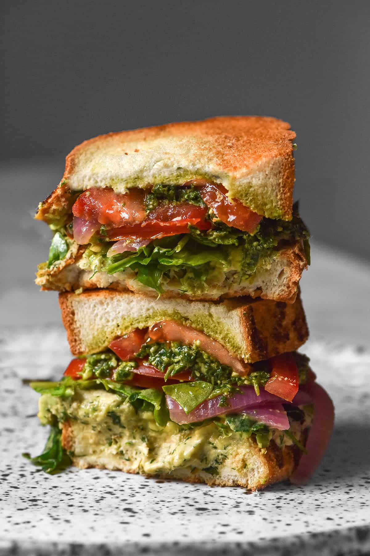 A side on close up image of a gluten free low FODMAP tunacado on a white speckled ceramic plate against a dark backdrop. The sandwich is filled with tuna mousse, rocket, pickled red onion, slices of tomato, avocado and low FODMAP pesto. The tunacado has been sliced and the pieces sit in a stack.