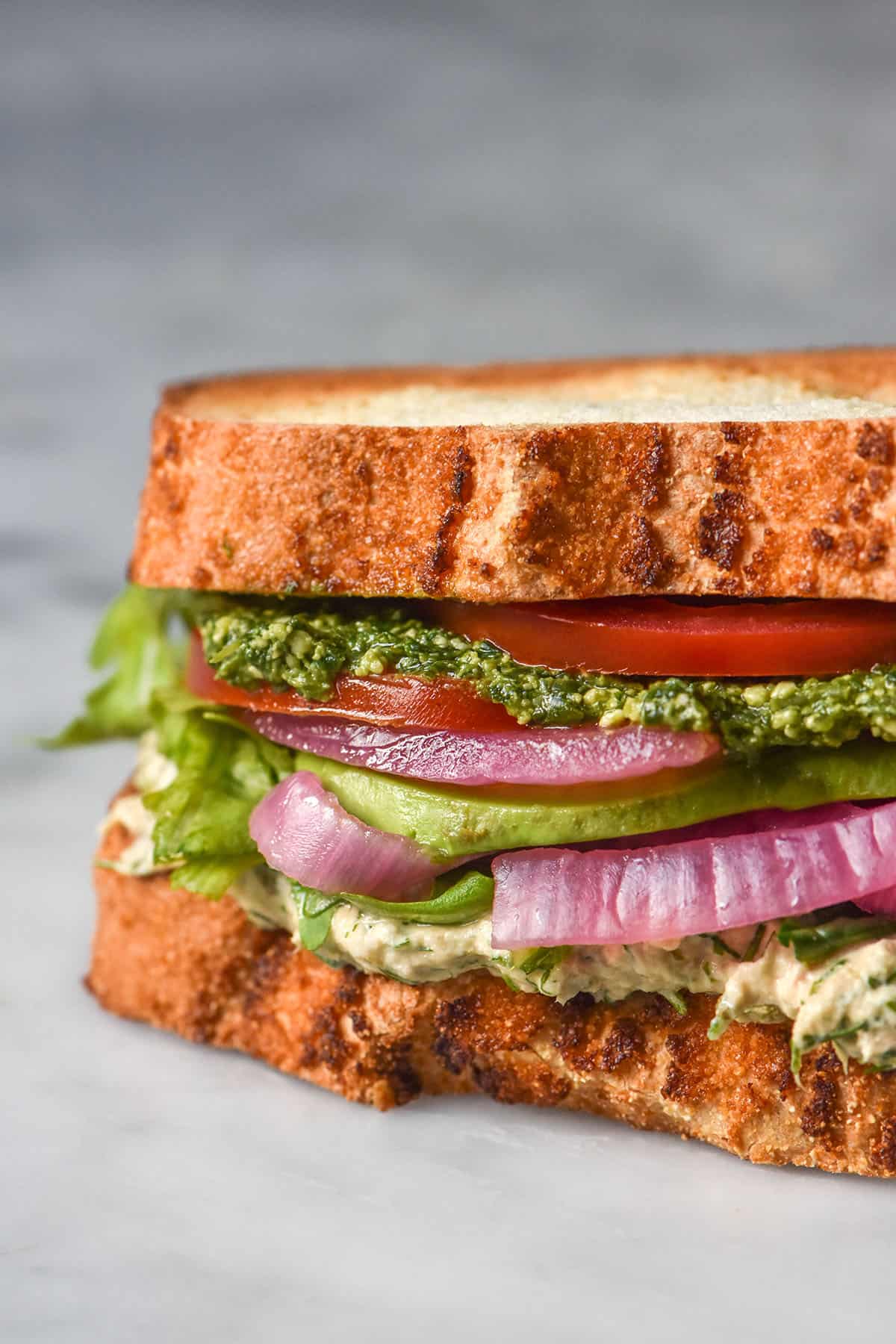 A side on close up image of a gluten free low FODMAP tunacado on a white marble table. The sandwich is filled with tuna mousse, rocket, pickled red onion, slices of tomato, avocado and low FODMAP pesto.