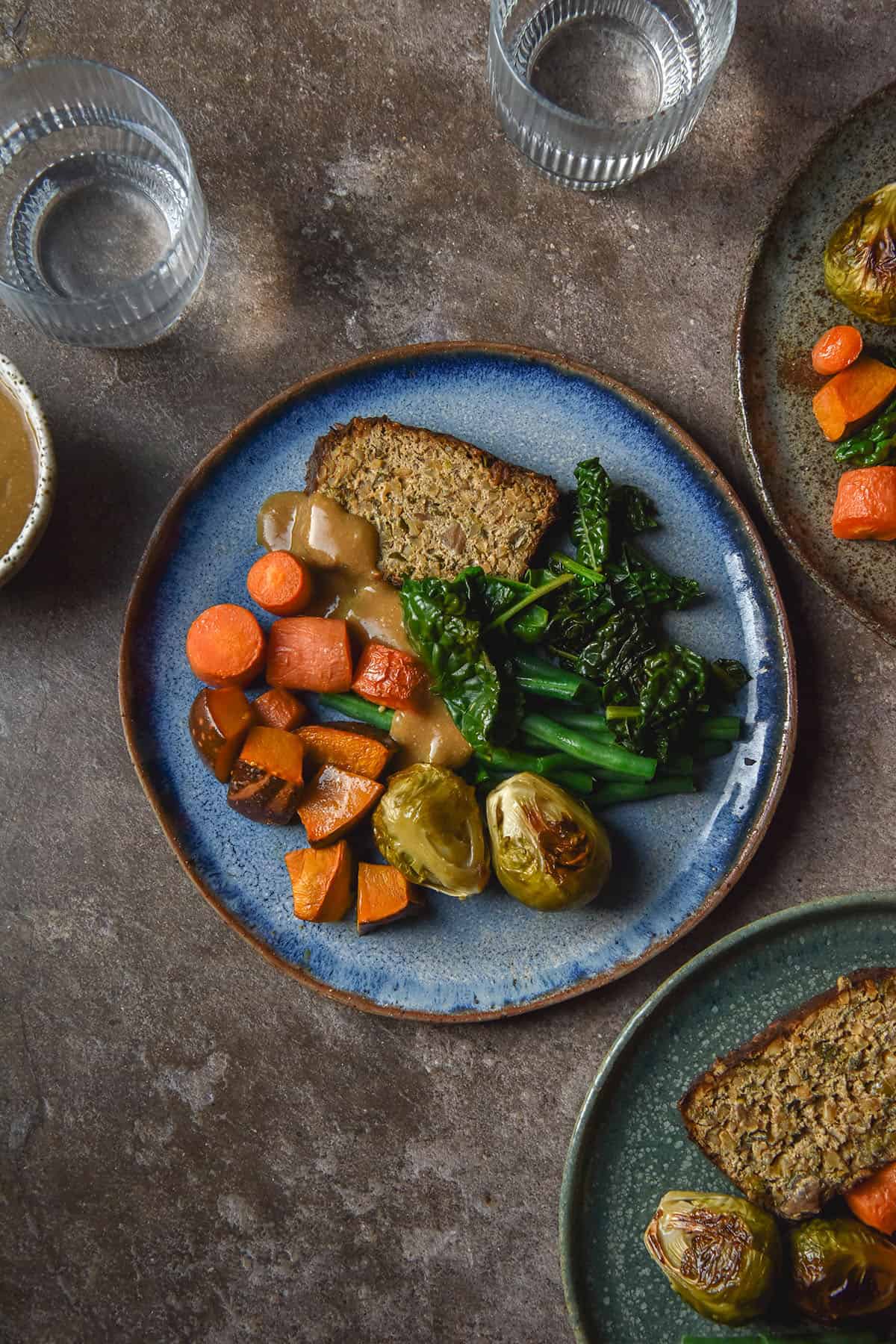 An aerial image of three plates topped with low FODMAP vegan meatloaf, roasted vegetables and gravy. The plates sit atop a dark grey backdrop and are surrounded by glasses of water