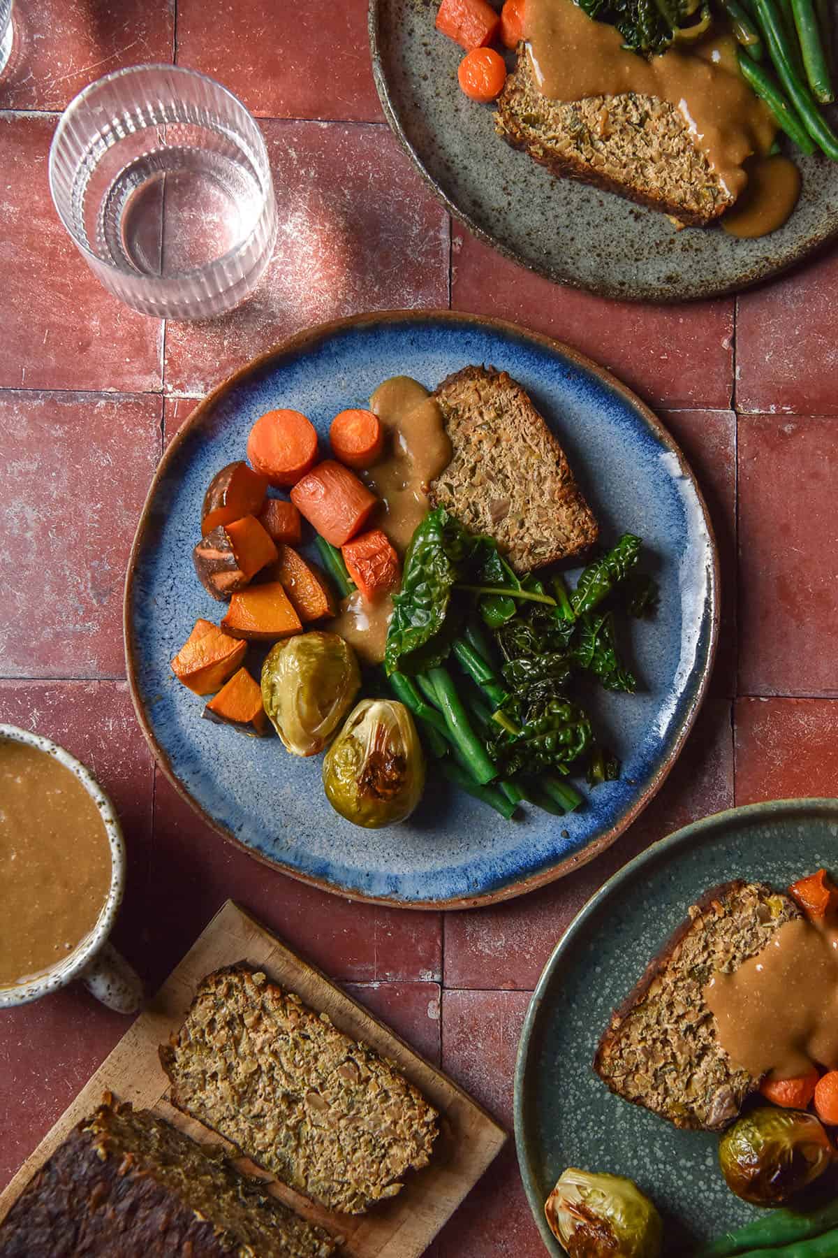 An aerial image of three plates topped with low FODMAP vegan meatloaf, roasted vegetables and vegan gravy. The plates sit atop a terracotta tile backdrop and extra nut loaf and gravy are casually arranged alongside the plates