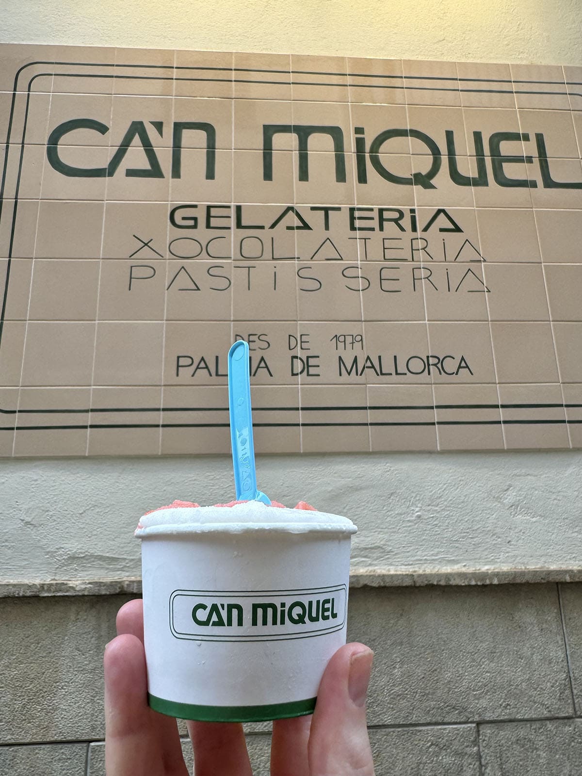 A photo of a cup of ice cream being up against the tiled sign of Can Miquel Heladeria in Palma, Mallorca