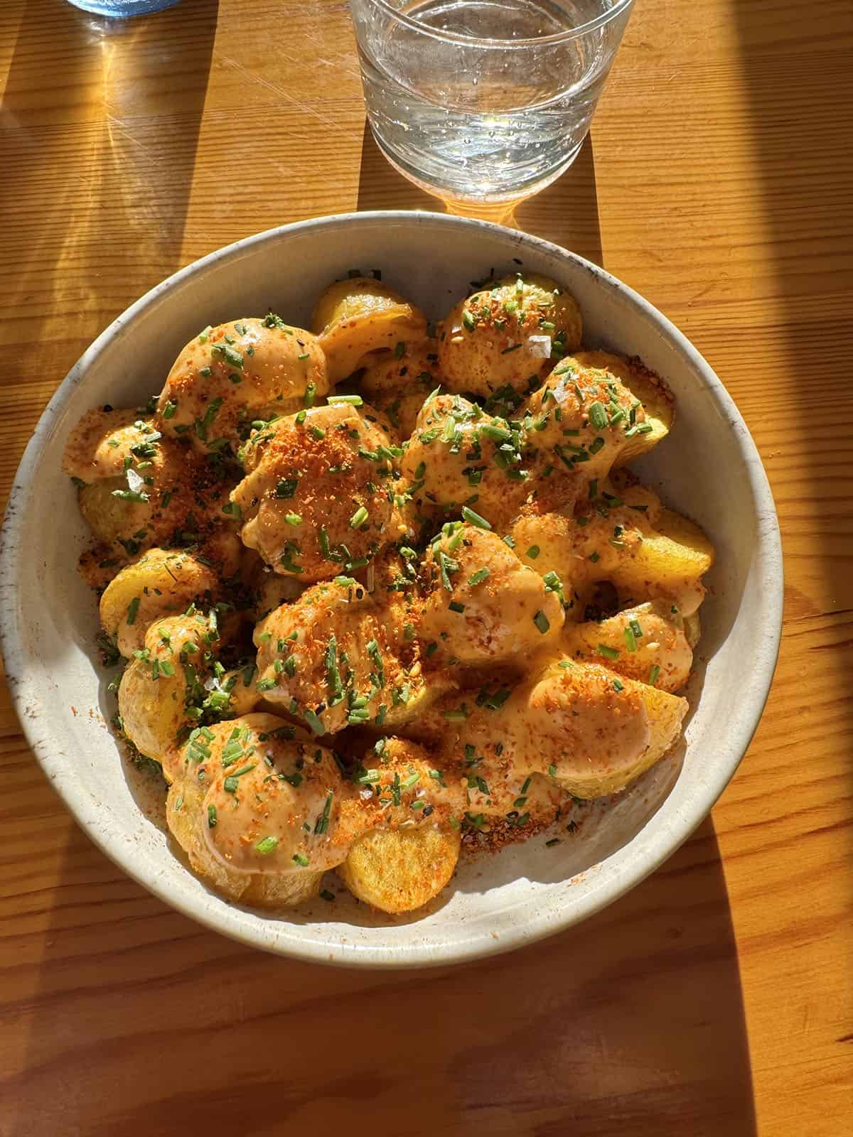 An aerial sunlit image of a bowl of patatas bravas and a glass of water on a wooden table