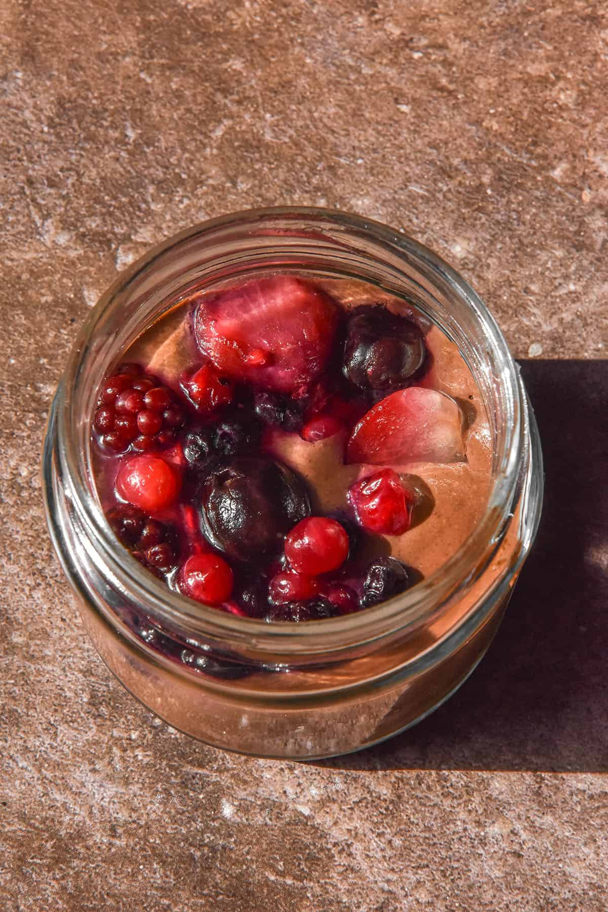 A brightly lit aerial image of a chocolate blended chia pudding topped with berries. The pudding is in a glass jar and sits atop a dark brown backdrop