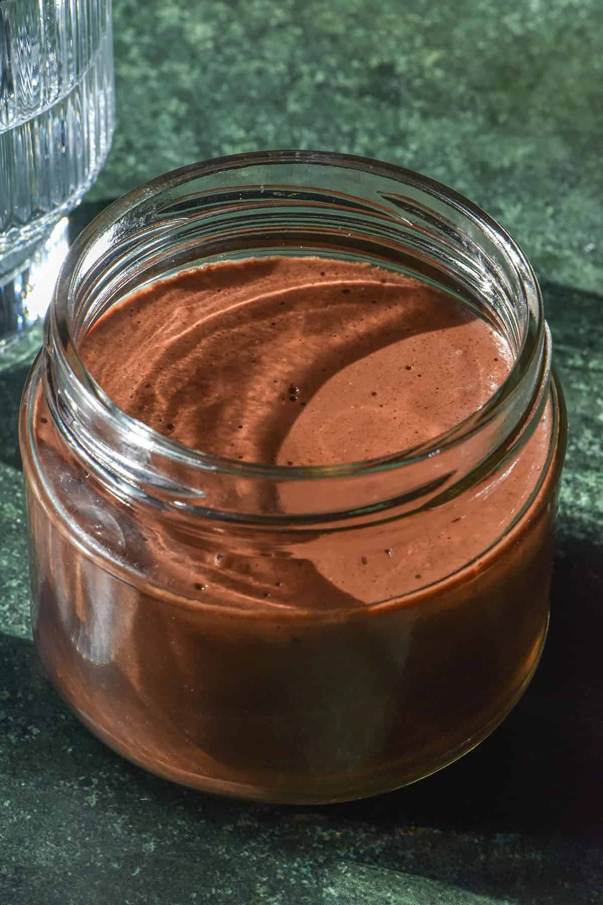 An aerial sunlit image of a blended chocolate chia pudding in a glass jar atop an olive green backdrop. The chia pudding is surrounded by sunlit glasses of water