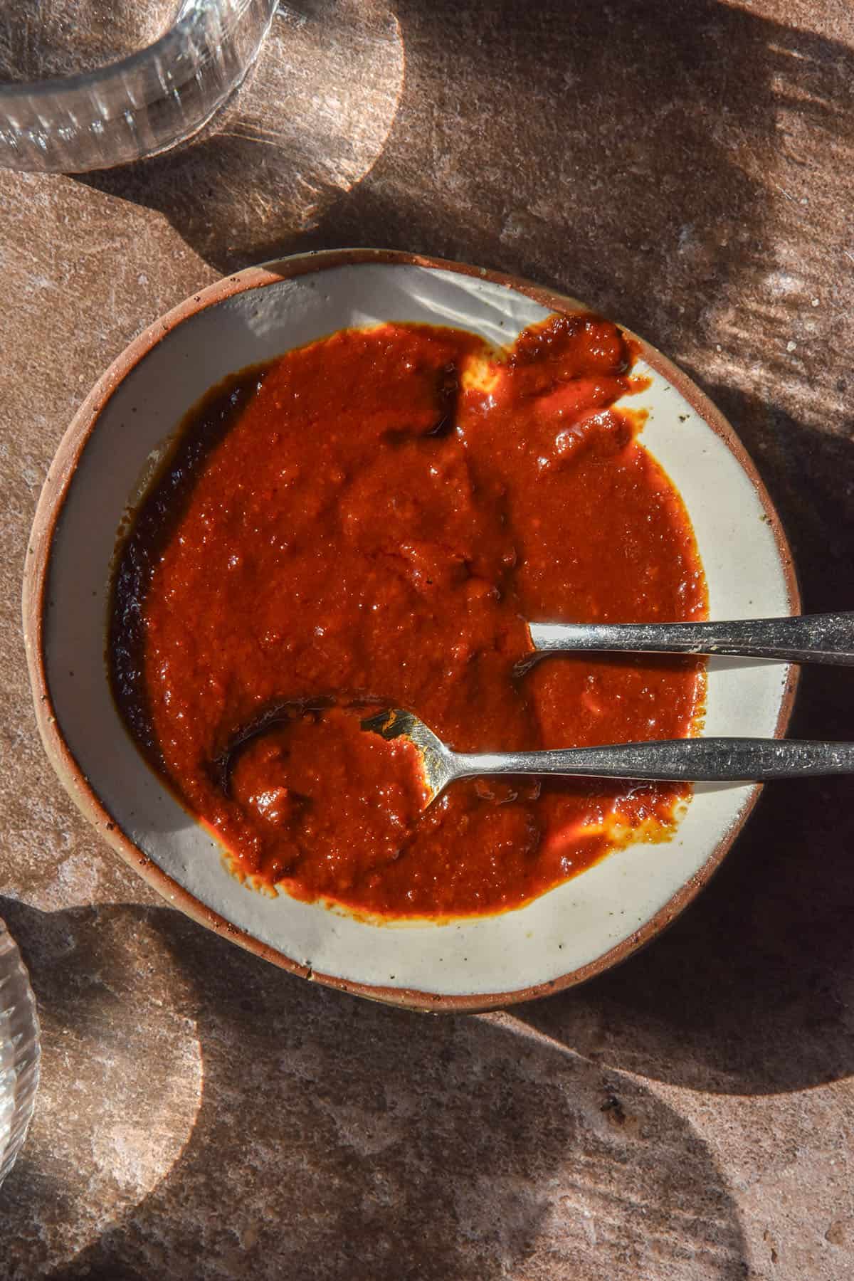 A sunlit aerial image of a white ceramic bowl filled with gluten free gochujang sauce. The bowl sits atop a dark brown backdrop with sunlit glasses of water surrounding it