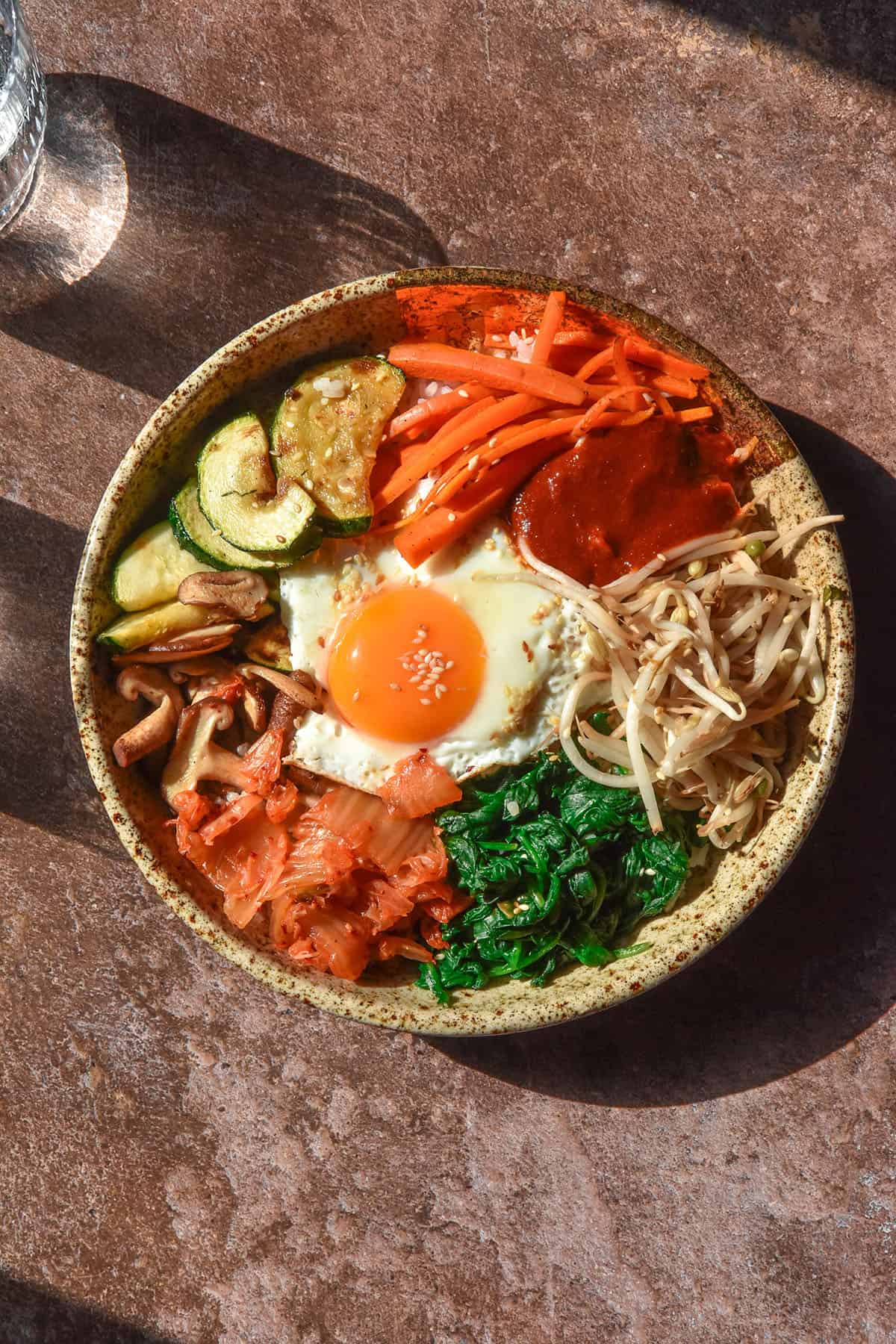 A sunlit aerial image of a bowl of gluten free vegetarian bibimbap. The bowl sits atop a dark brown backdrop surrounded by sunlit glasses of water