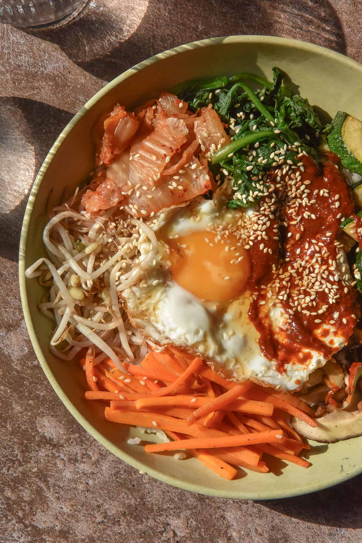 A sunlit aerial image of a bowl of vegetarian gluten free bibimbap atop a dark brown backdrop