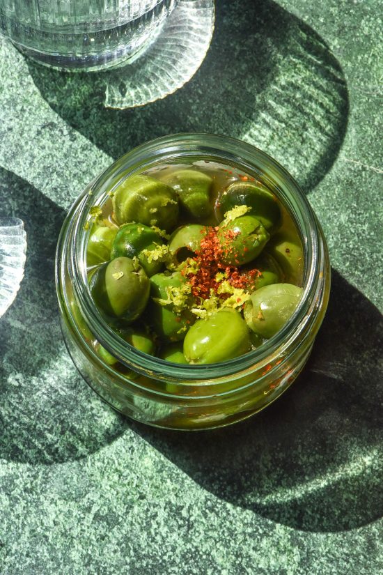 A sunlit aerial image of a jar of Italian margarita olives on an olive green backdrop in bright sunlight. Two sunlit glasses of water sit to the left of the olives.