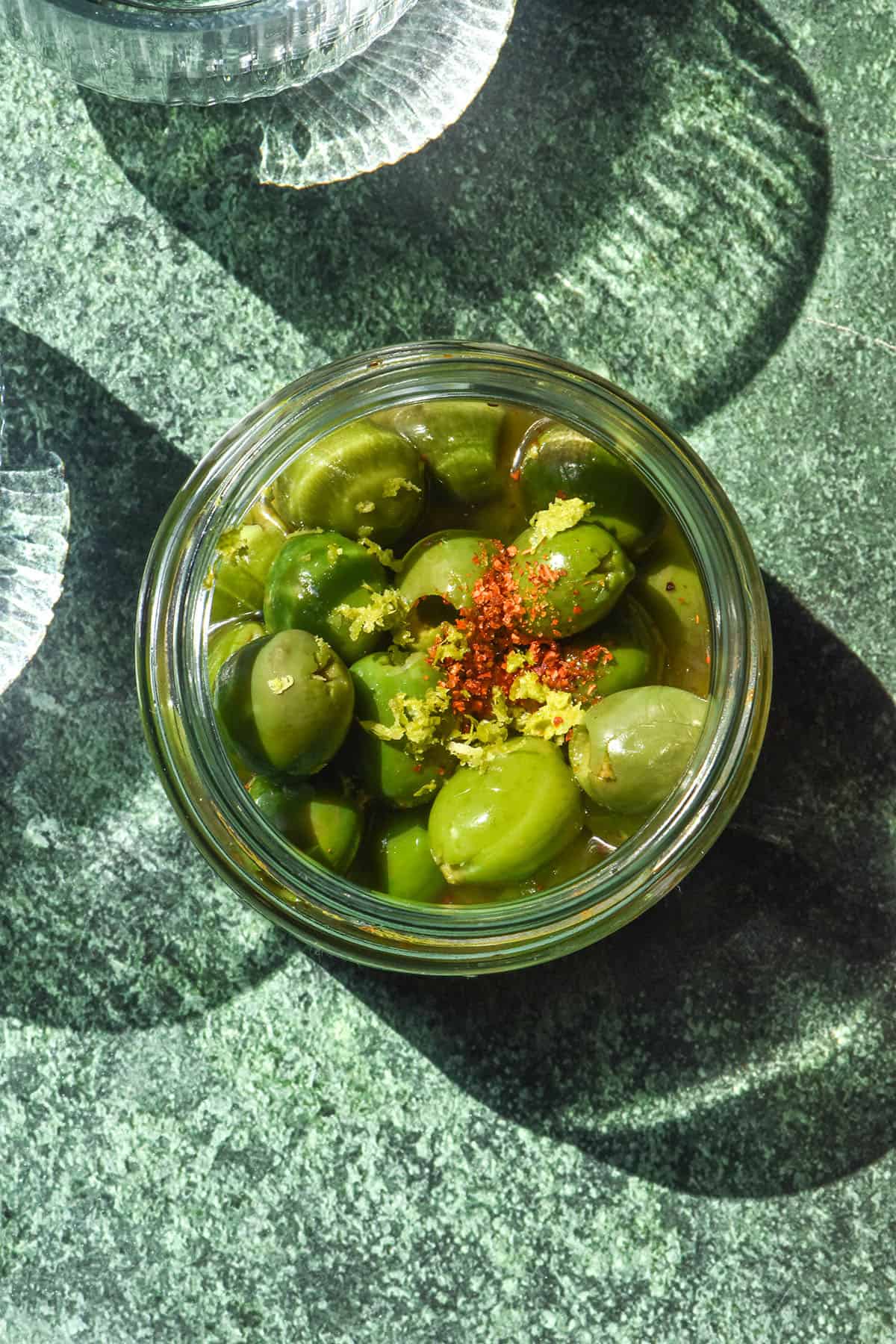 A sunlit aerial image of a jar of Italian margarita olives on an olive green backdrop in bright sunlight. Two sunlit glasses of water sit to the left of the olives.