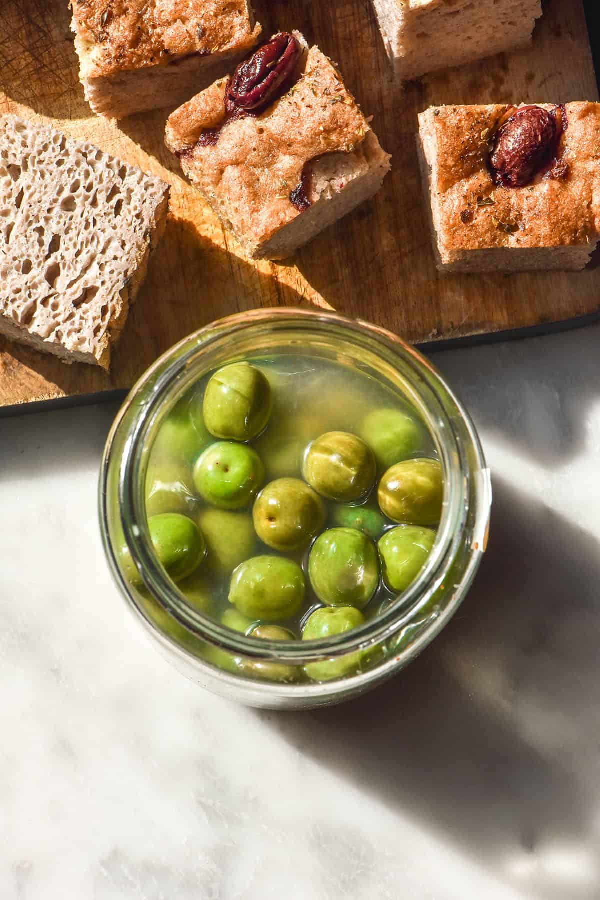 An aerial image of a jar of Italian margarita olives on a white marble table in bright sunlight. A wooden board of chopped gluten free focaccia sits above the jar of olives.