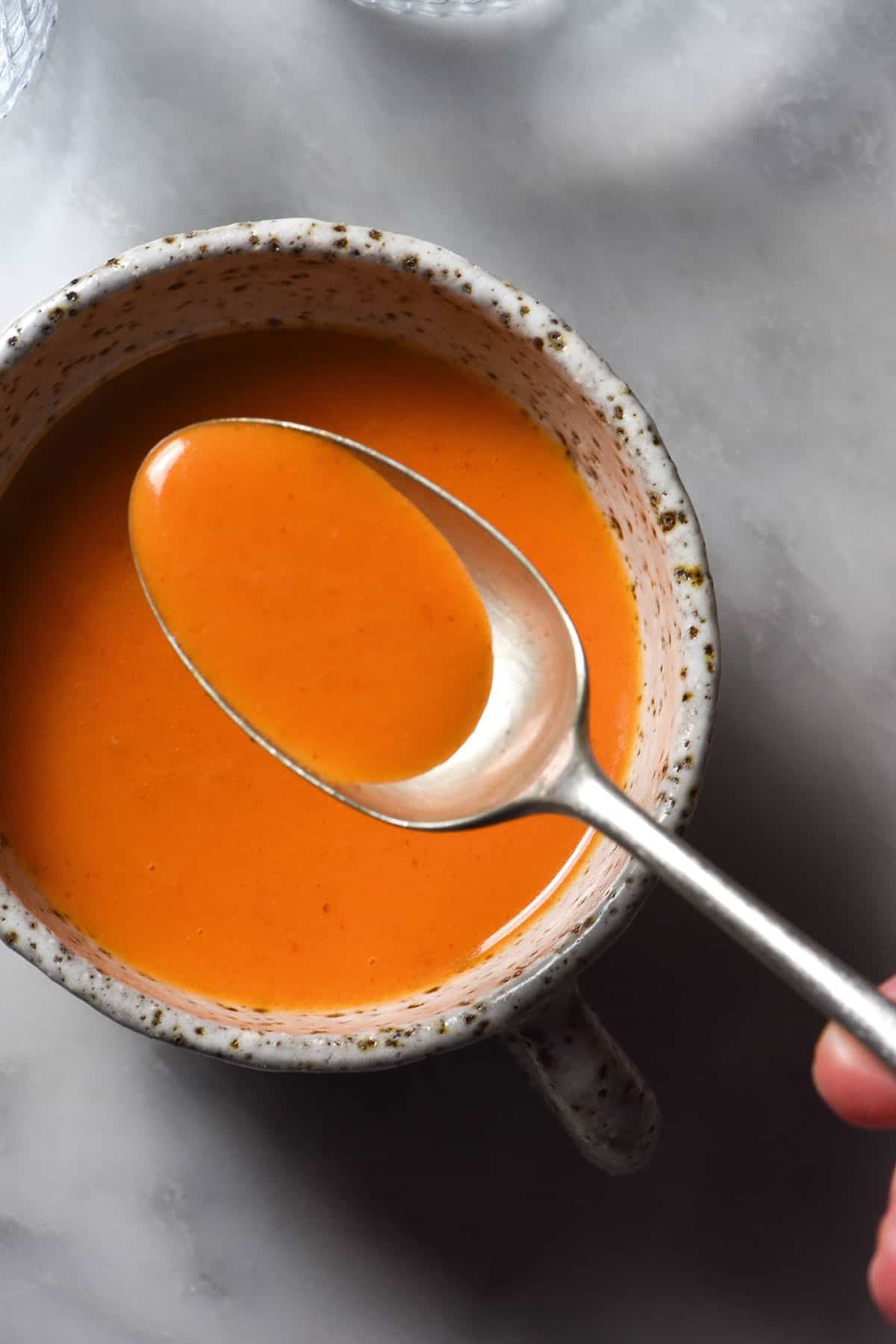 An aerial sunlit image of white ceramic mug filled with low FODMAP Buffalo sauce on a white marble table. A spoon dips into the Buffalo sauce