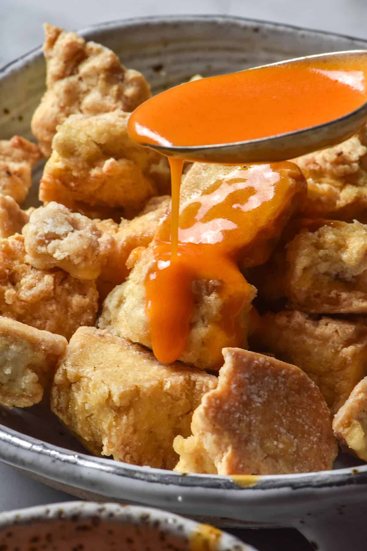 A side on close up of some tofu nuggets being drizzled with spoonfuls of low FODMAP Buffalo sauce