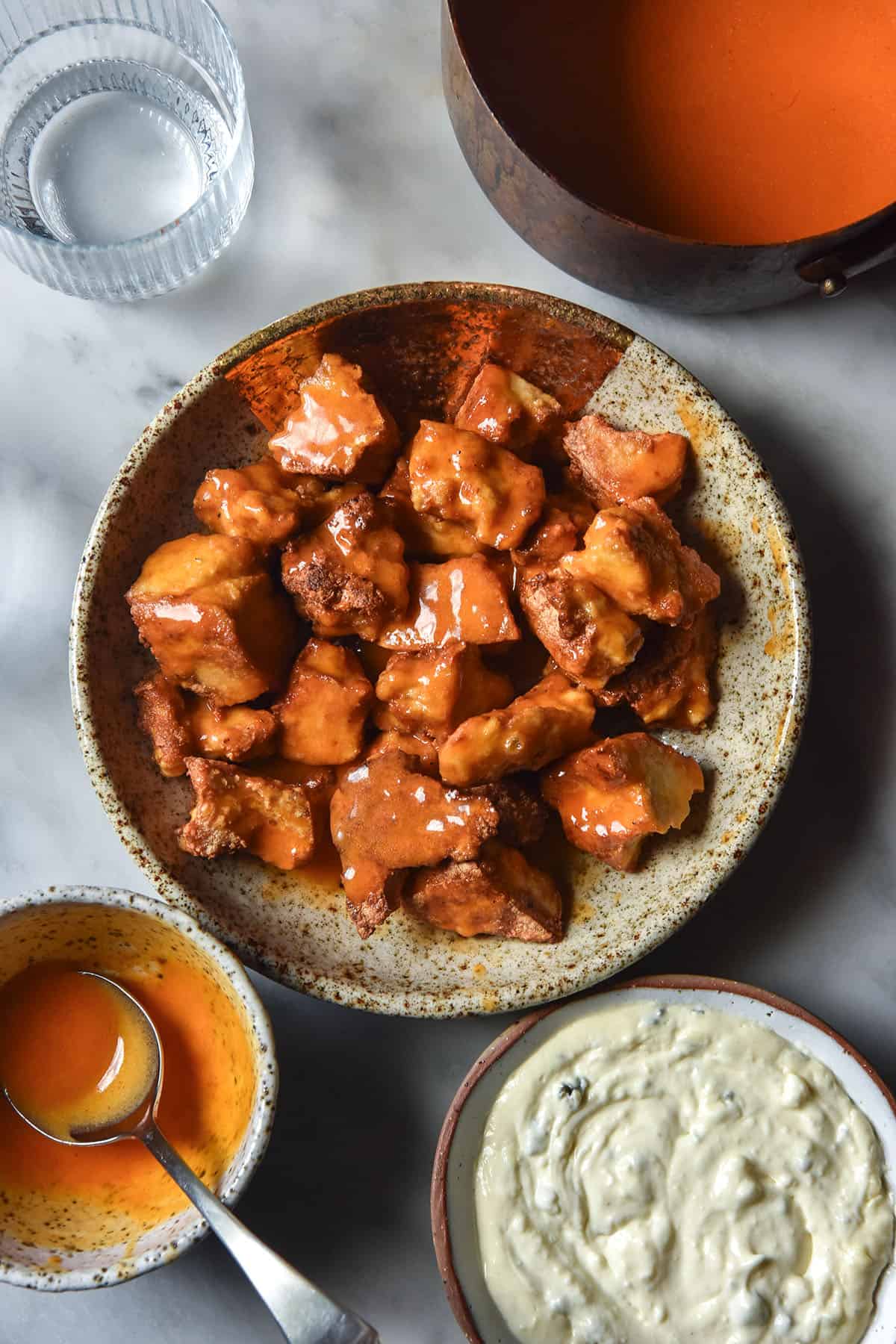 An aerial image of a ceramic beige bowl filled with low FODMAP Buffalo tofu, surrounded by cooking vessels filled with Buffalo sauce, a water glass and a small bowl of low FODMAP blue cheese dip all on a white marble table