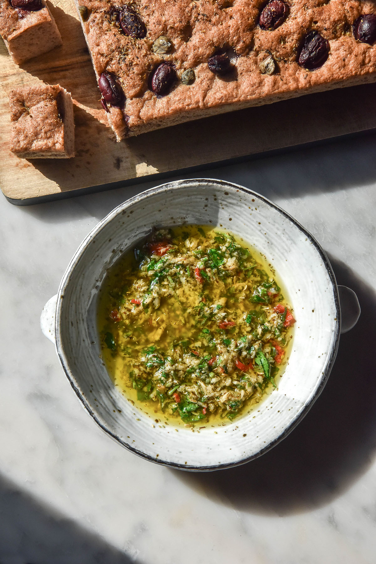 An aerial image of a white ceramic bowl filled with low FODMAP bread dipping oil atop a white marble table. A wooden chopping board topped with a gluten free olive focaccia sits above the bowl of dipping oil.