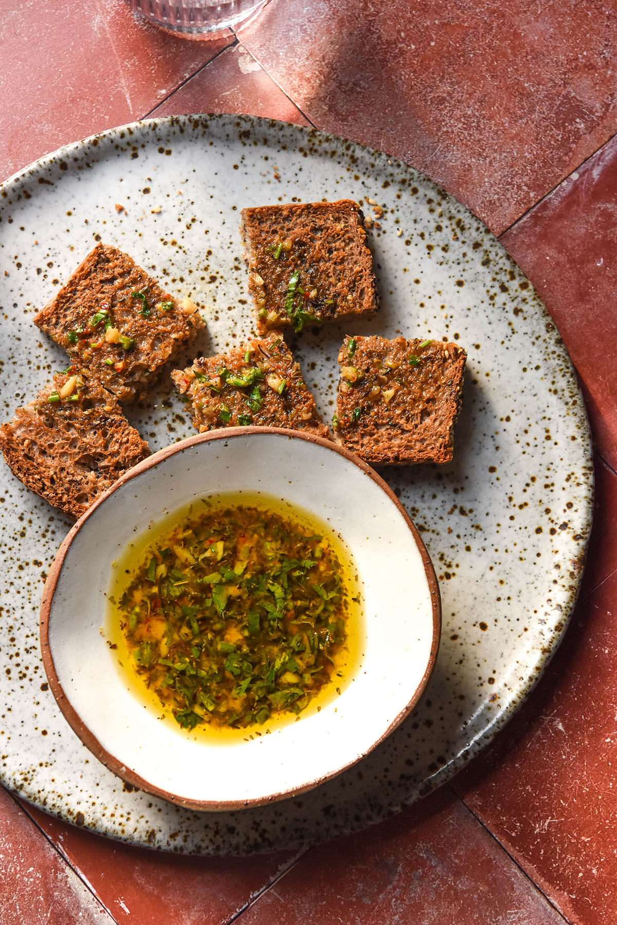 An aerial image of low FODMAP bread dipping oil in a small white bowl atop a speckled ceramic plate. The bowl is surrounded by toasted buckwheat bread and the plate sits atop a terracotta tile backdrop