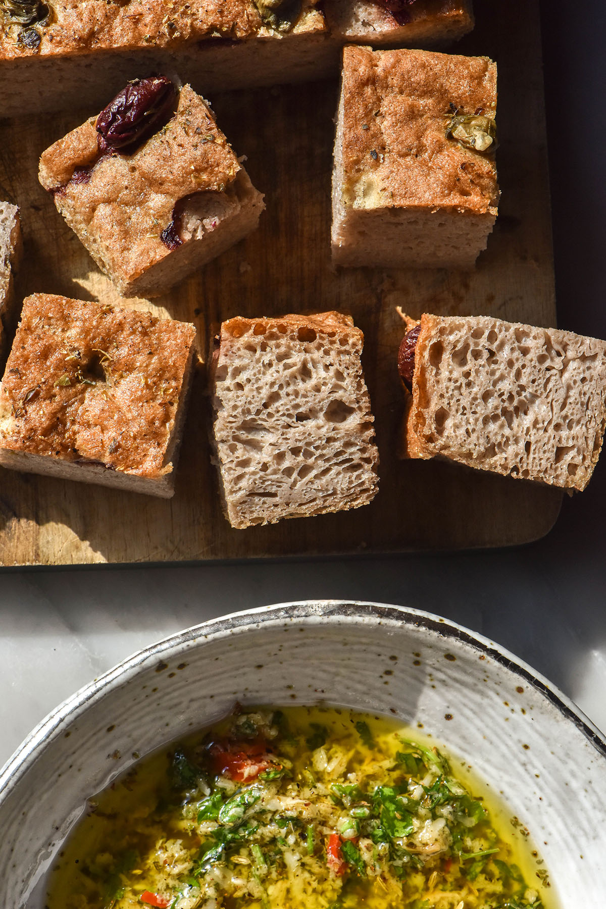 An aerial image of a white ceramic bowl filled with low FODMAP bread dipping oil underneath a wooden chopping board with pieces of buckwheat focaccia