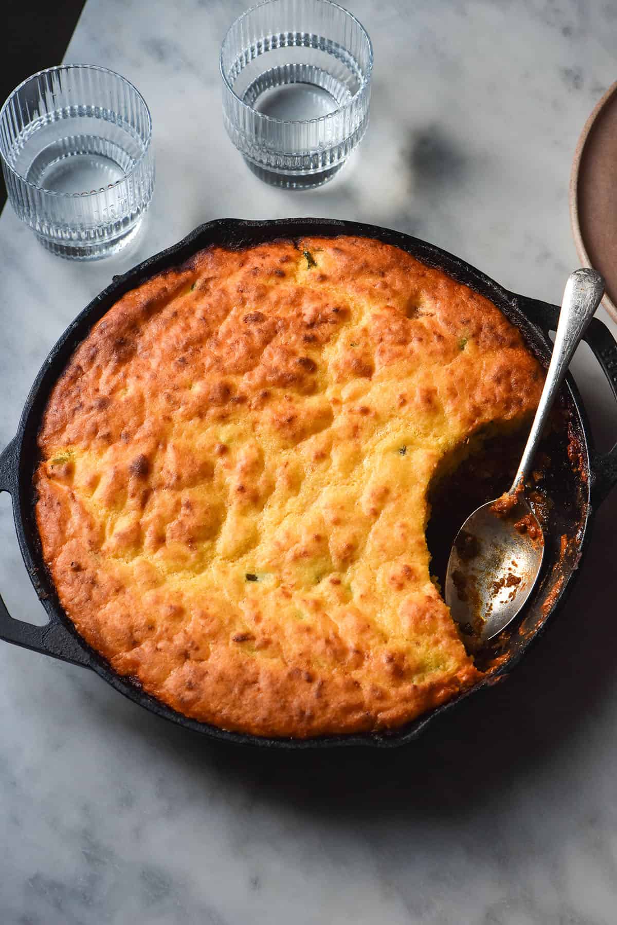 An aerial image of a low FODMAP vegetarian cornbread chilli in a black skillet atop a white marble table. Two glasses of water sit to the top of the cornbread