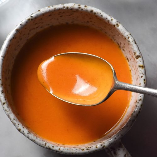 An aerial sunlit image of white ceramic mug filled with low FODMAP Buffalo sauce on a white marble table. A spoon dips into the Buffalo sauce