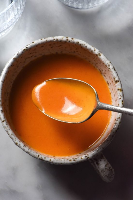 An aerial sunlit image of white ceramic mug filled with low FODMAP Buffalo sauce on a white marble table. A spoon dips into the Buffalo sauce