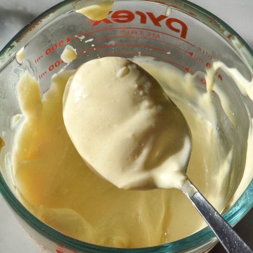 An aerial sunlit image of a jug of low FODMAP mayonnaise atop a white marble table in bright sunlight. A spoon sits on the edge of the jug with a spoonful of mayonnaise