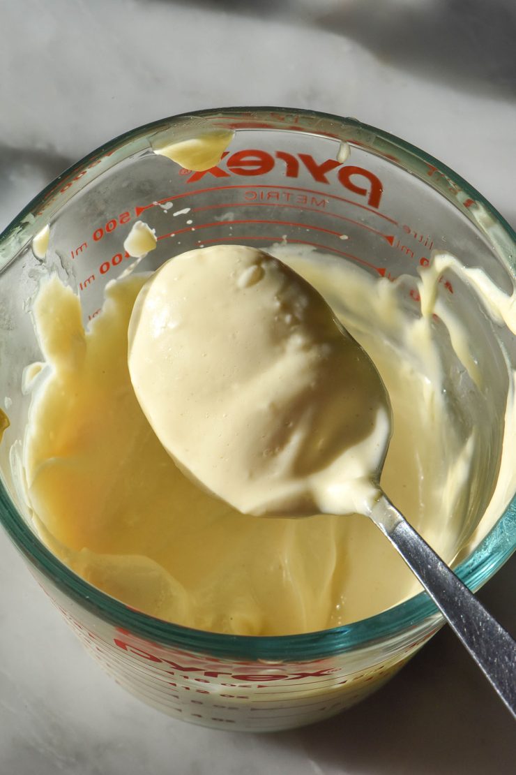 An aerial sunlit image of a jug of low FODMAP mayonnaise atop a white marble table in bright sunlight. A spoon sits on the edge of the jug with a spoonful of mayonnaise