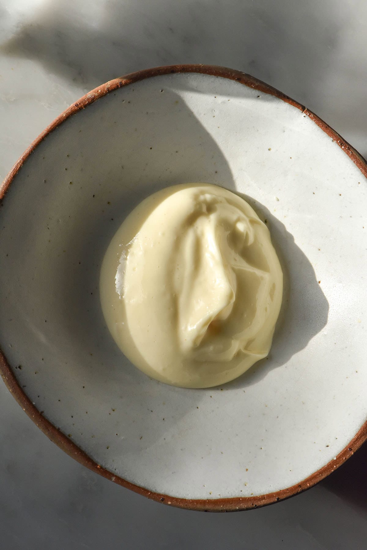 An aerial image of a dollop of low FODMAP mayonnaise in a white ceramic bowl atop a sunlit white marble table