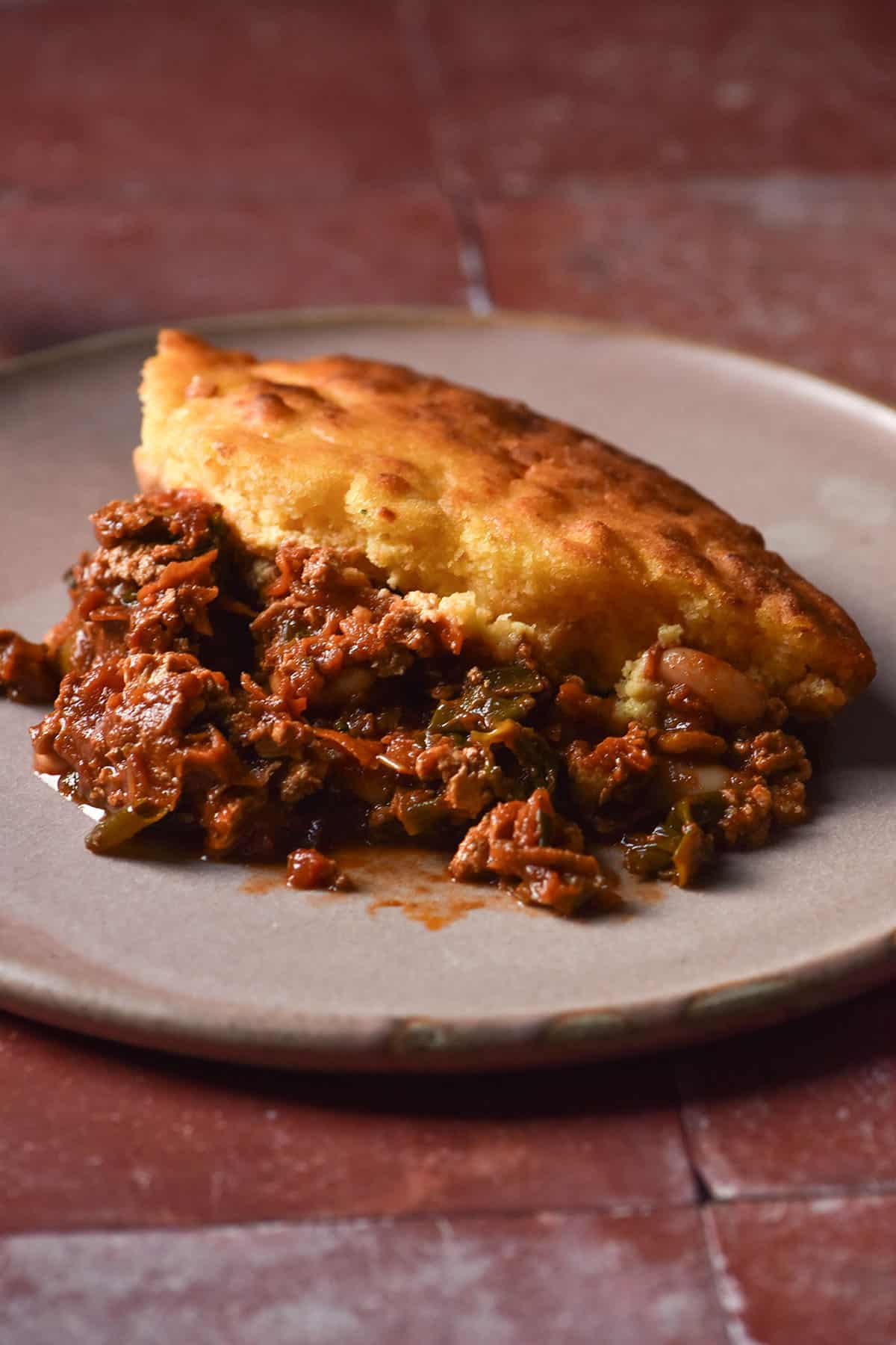 A side on image of a pink ceramic plate topped with low FODMAP cornbread chilli. The plate sits atop a terracotta tile backdrop