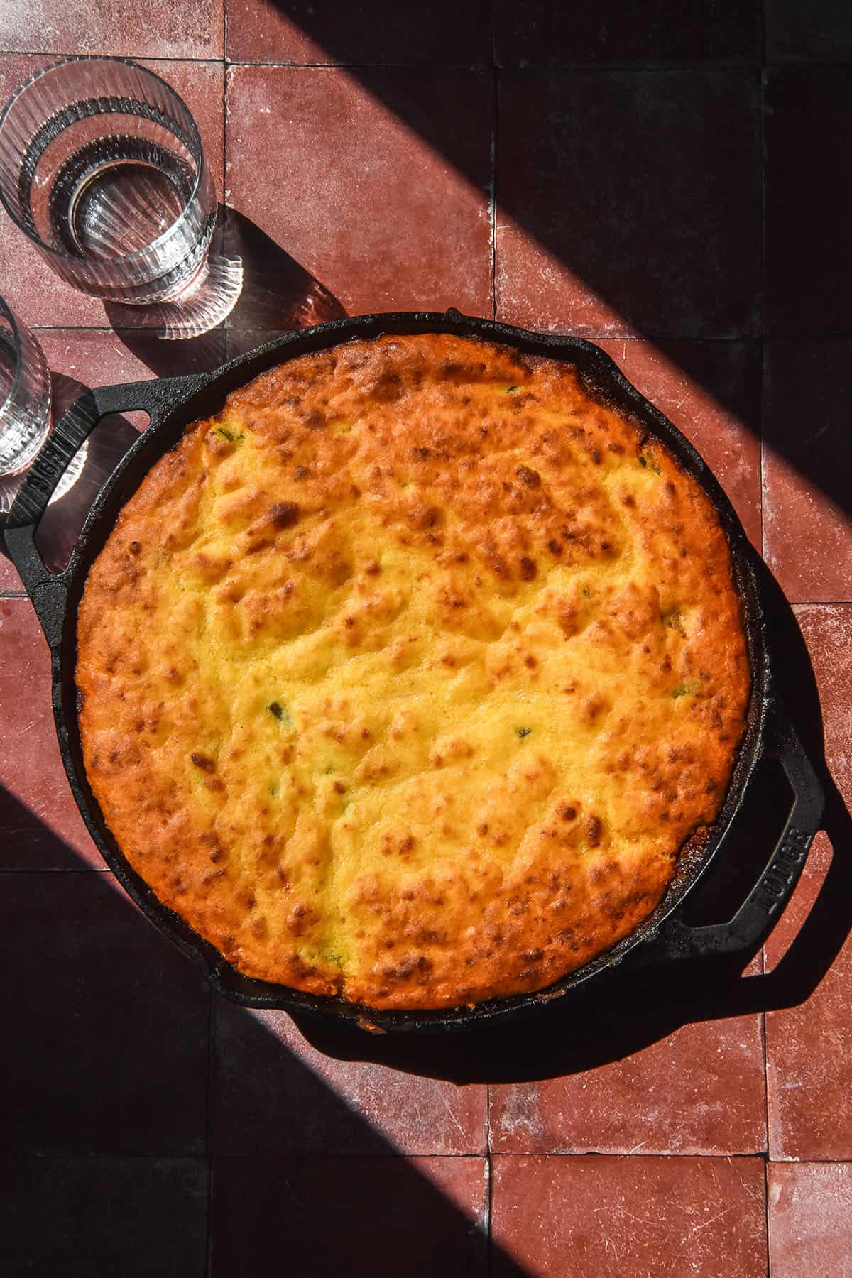 An aerial image of a low FODMAP vegetarian cornbread chilli in a black skillet. The cornbread sits atop a terracotta tile backdrop in contrasting sunlight. Two sunlit water glasses sit to the left of the cornbread