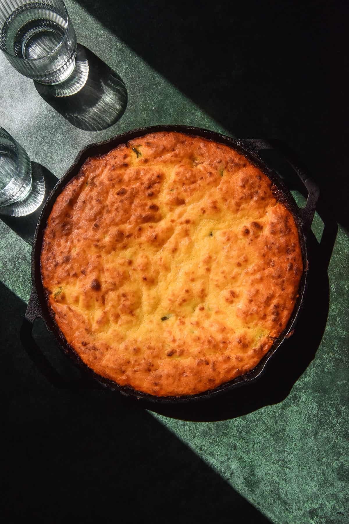 A sunlit aerial image of a low FODMAP vegetarian cornbread chilli in a black skillet atop an olive green backdrop. The skillet sits in contrasting sunlight and two sunlit glasses of water sit to the top left of the skillet