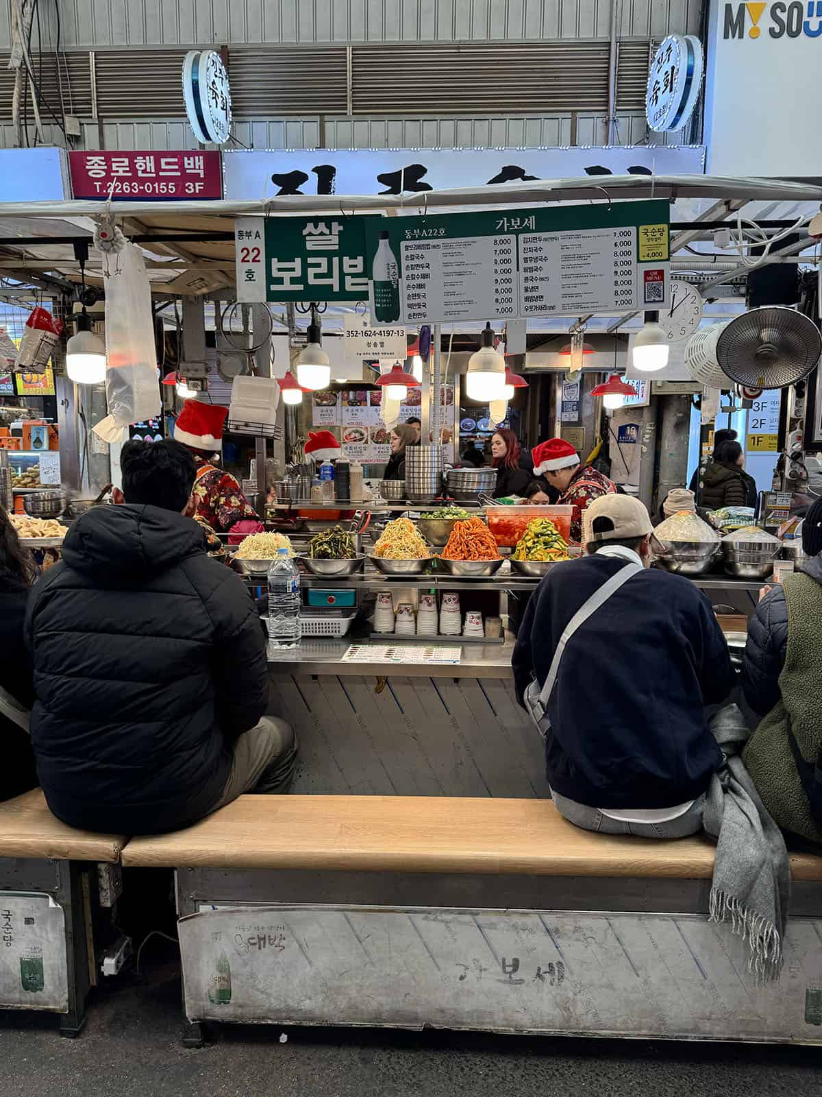 An image of a bibimbap stall at Gwanjang market in Seoul, South Korea. The ladies serving the bibimbap are wearing Santa hats