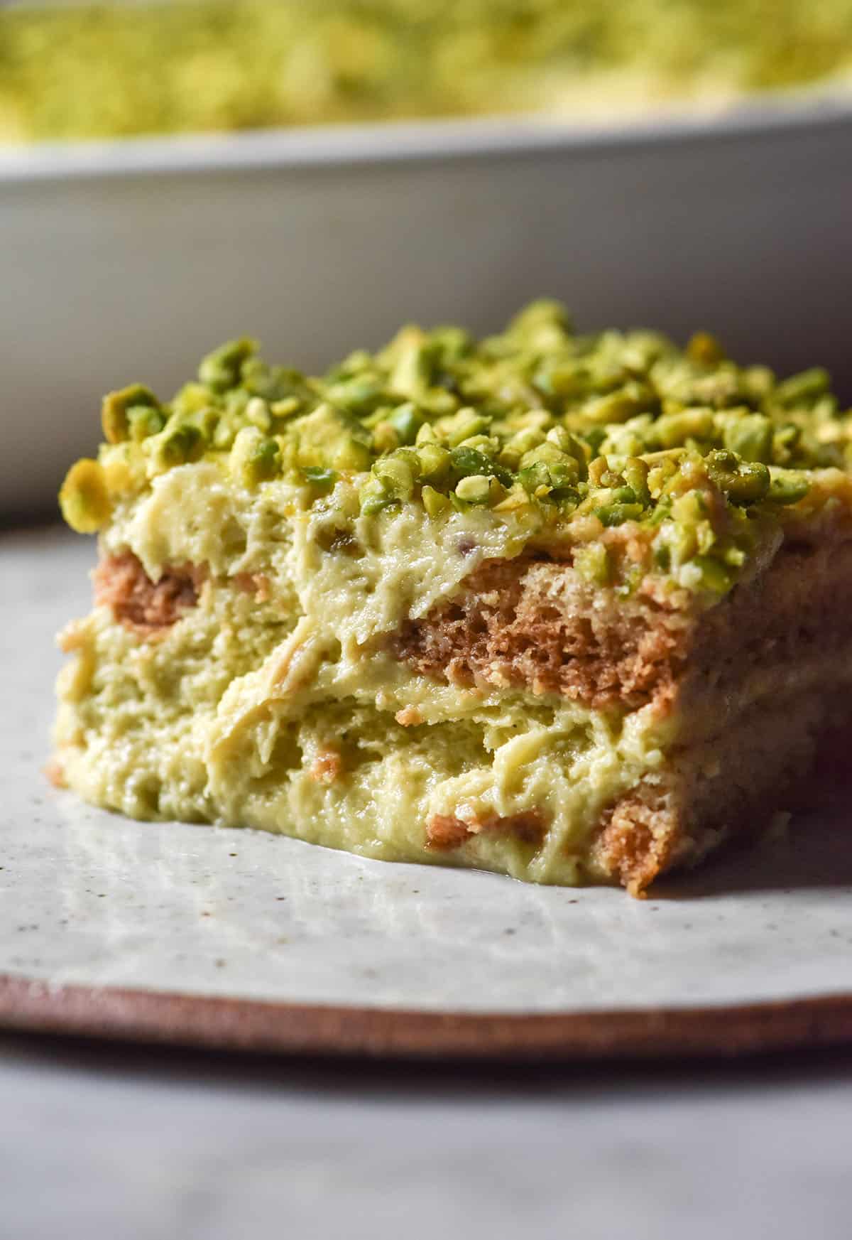 A side on image of a slice of gluten free Pistachio Tiramisu on a white speckled ceramic plate atop a white marble table. The white ceramic dish that holds the remaining Tiramisu sits in the background