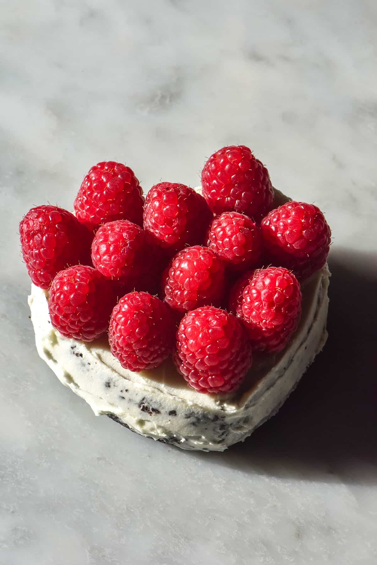 A sunlit aerial image of a mini gluten free chocolate cake topped with vanilla icing and raspberries. The cake is in the shape of a love heart and sits atop a white marble table