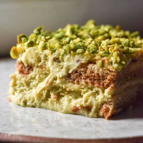 A side on image of a slice of gluten free Pistachio Tiramisu on a white speckled ceramic plate atop a white marble table. The white ceramic dish that holds the remaining Tiramisu sits in the background