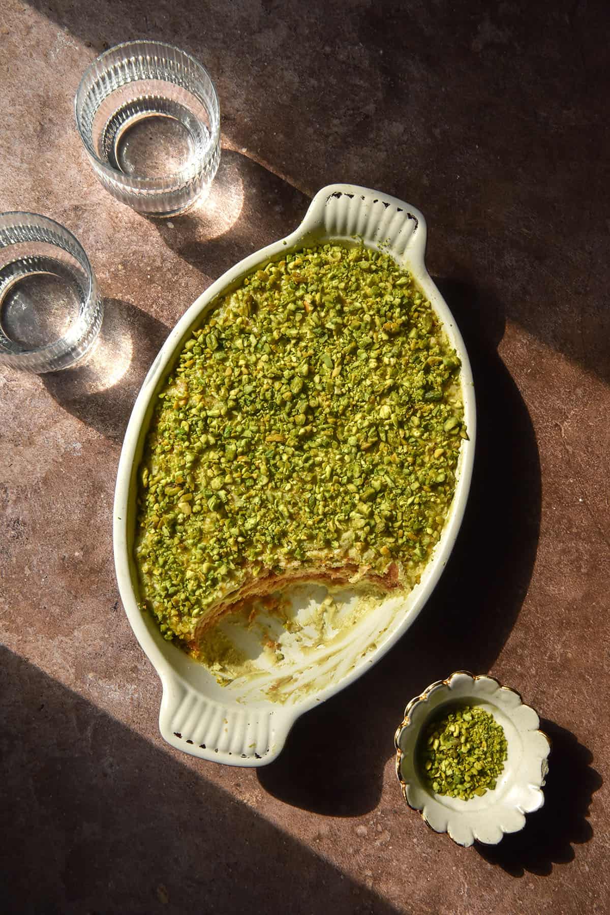 An aerial image of a gluten free pistachio Tiramisu in a white ceramic dish atop a dark grey background in contrasting sunlight. Two sunlit water glasses sit to the left of the Tiramisu and a small ceramic bowl of chopped pistachios sits to the right of the image.