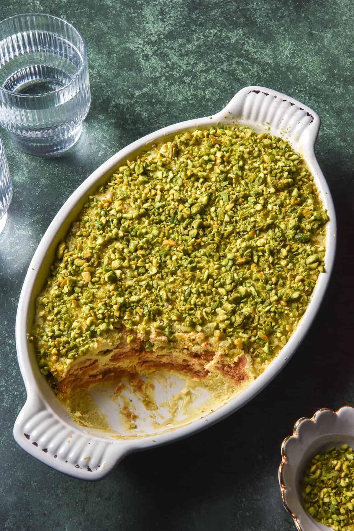 An aerial image of a gluten free pistachio Tiramisu in a white ceramic dish. The Tiramisu sits atop an olive green backdrop. Two glasses of water sit to the left of the Tiramisu and a small ceramic bowl of chopped pistachios sits to the right of the Tiramisu. 