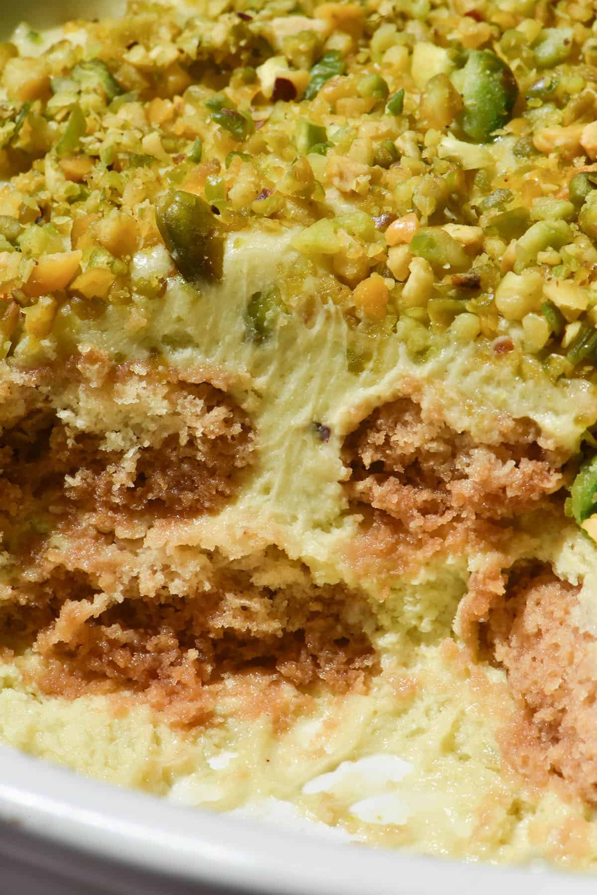 A macro close up image of a pistachio tiramisu topped with crushed pistachios in a white ceramic plate. A slice has been taken from the tiramisu, revealing the gluten free savoiardi inside.