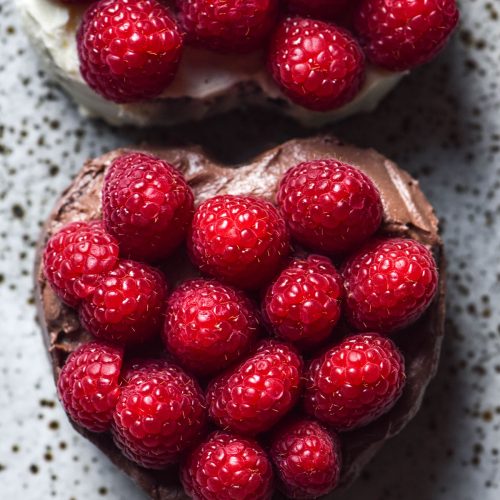 An aerial sunlit image two mini heart shaped gluten free chocolate cakes topped with raspberries